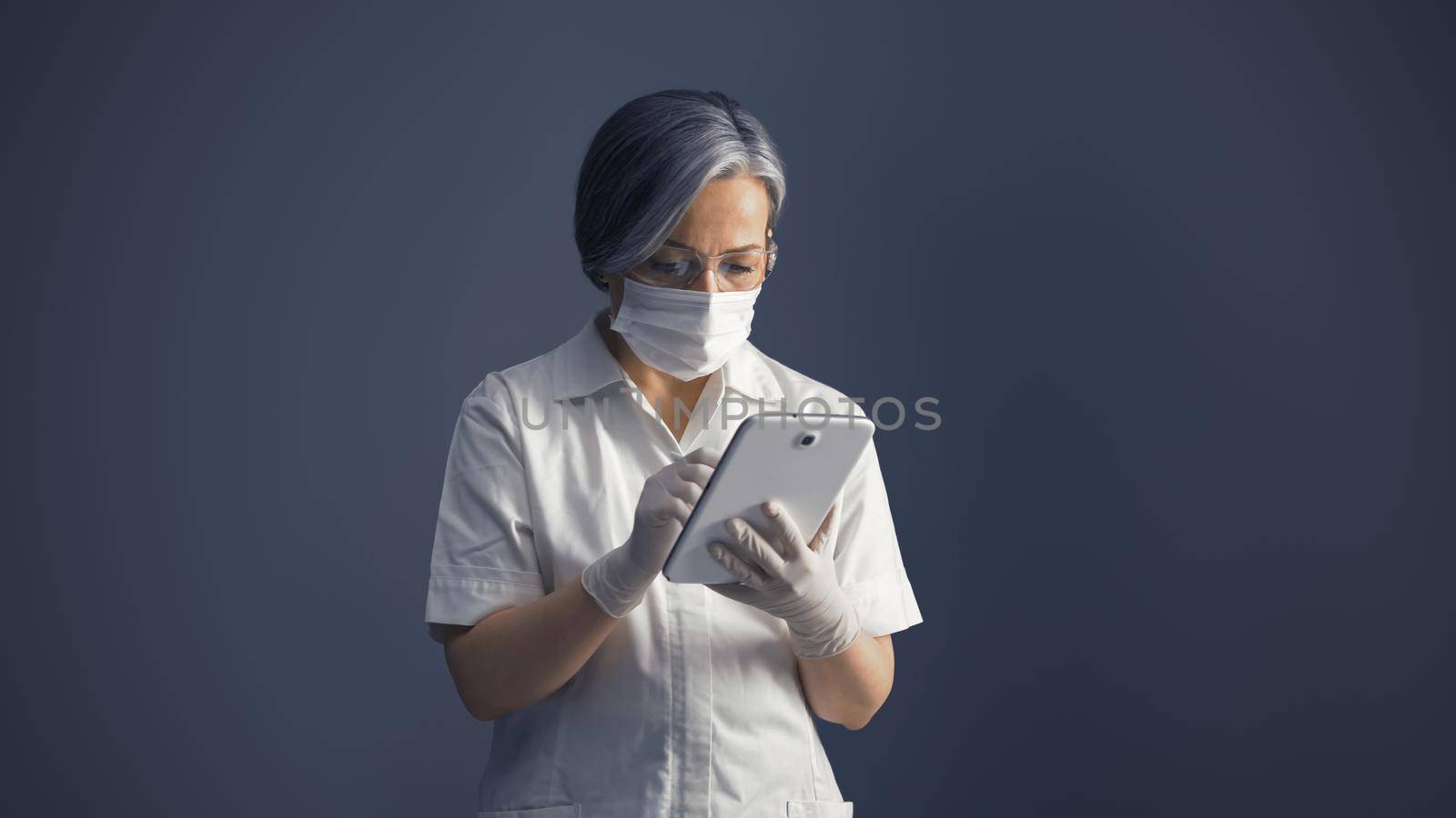 Masked female doctor using digital tablet. Serious Gray haired woman working with digital app or data base by LipikStockMedia