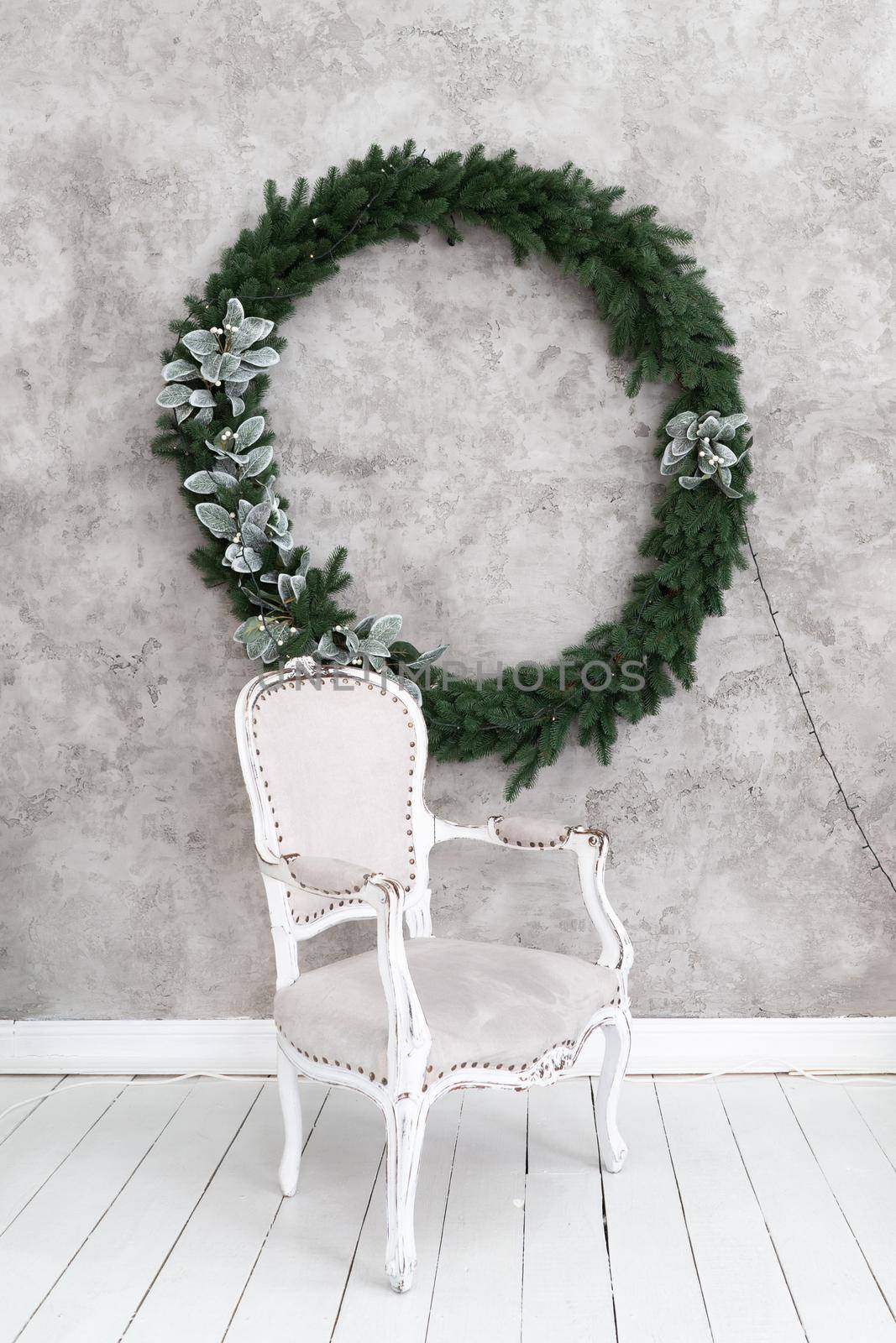 Christmas interior of the room. Gray chair stands under a light wall on which hangs a Christmas wreath. Green conifer wreath decorated with silver leaves and garland. High quality photo.
