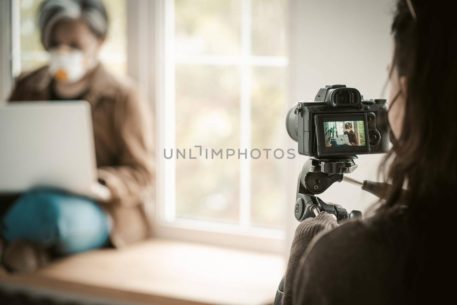 Backstage of shooting. Selective focus on digital camera in foreground. Woman in face mask using laptop sitting near window on blurred background. Quarantine concept.