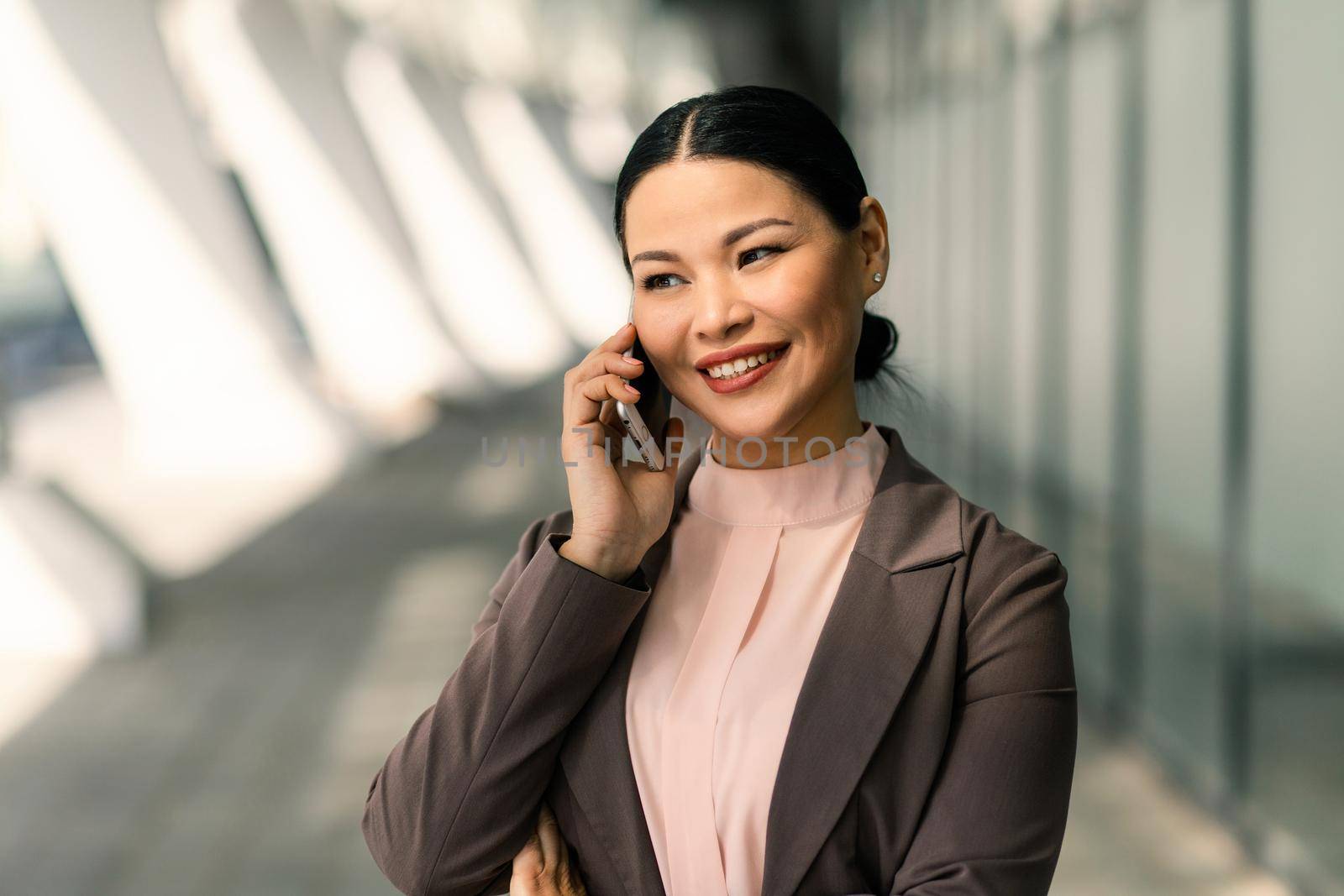 Smiling Asian businesswoman in suit talking on mobile phone against backdrop of business center. High quality photo.