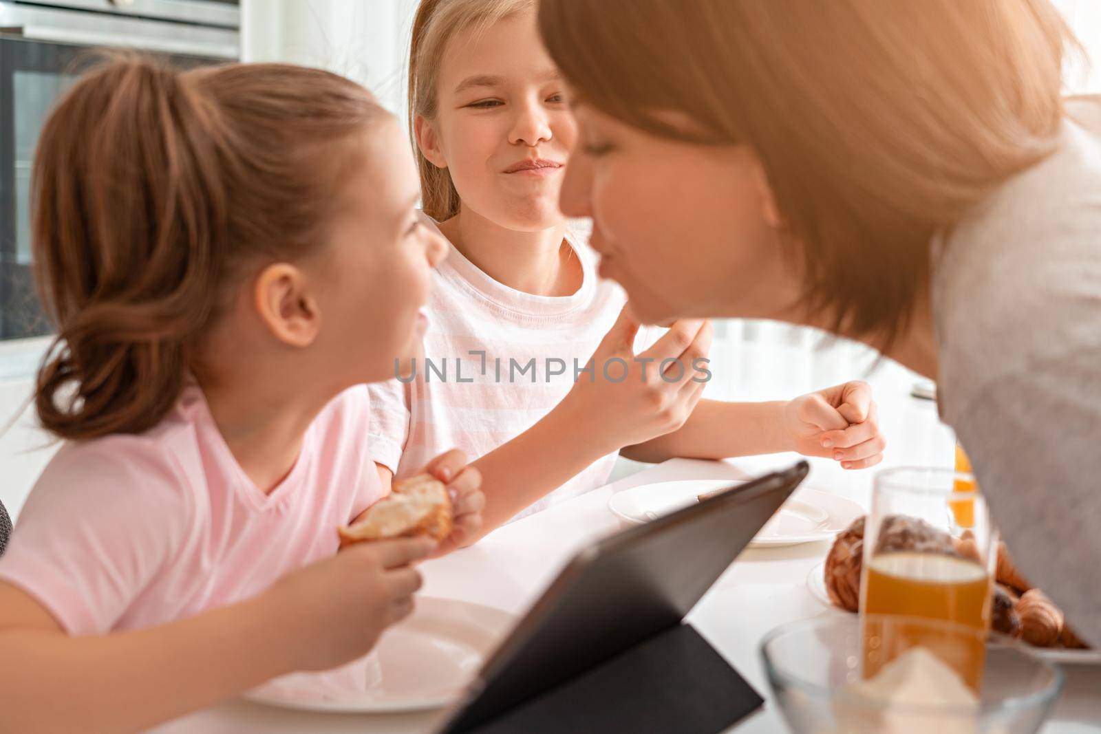 Mother trying to kiss daughter while having breakfast together with two kids by Mariakray