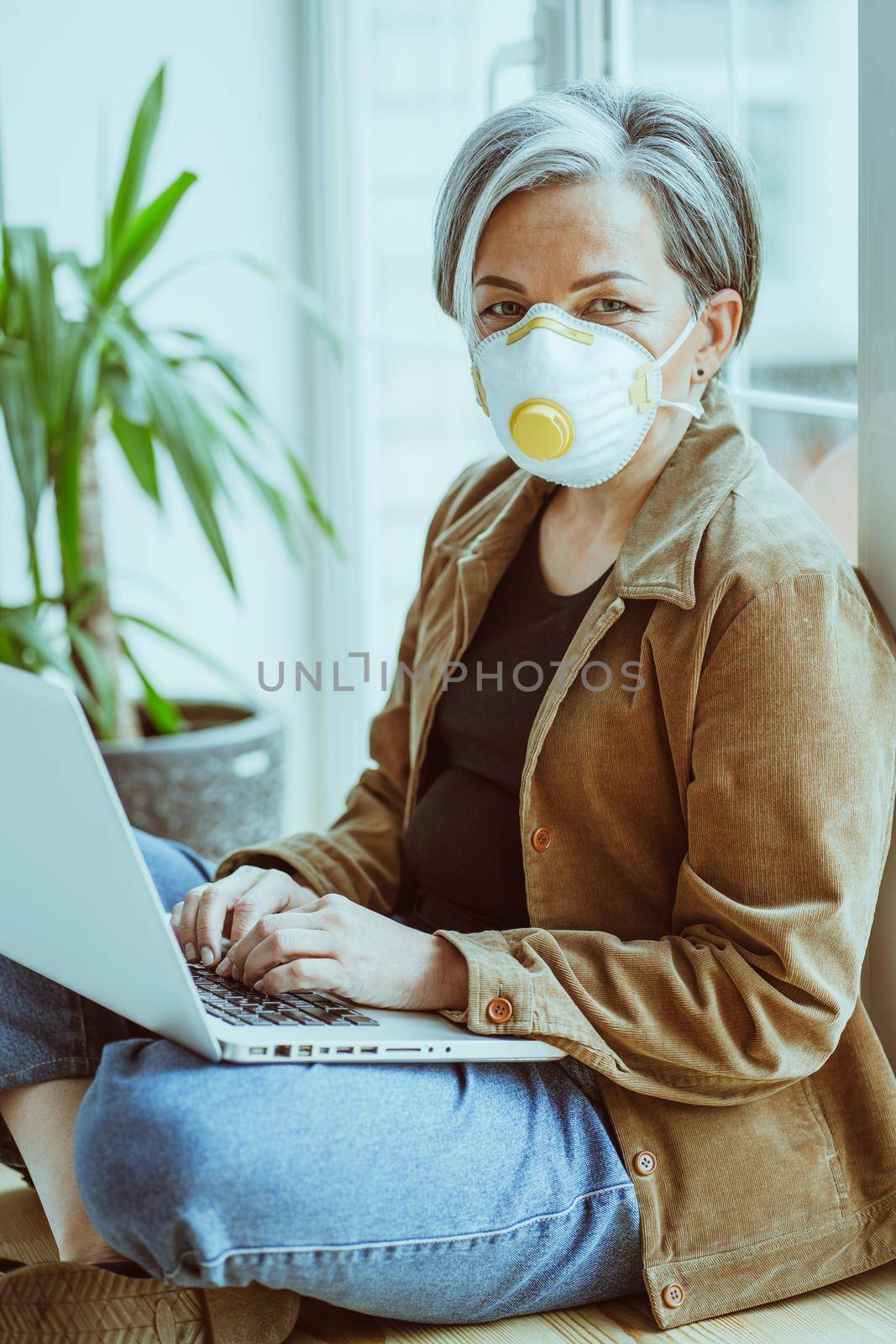 Masked aged woman looks camera using laptop while sitting crossed legs on windowsill. Pandemic concept.