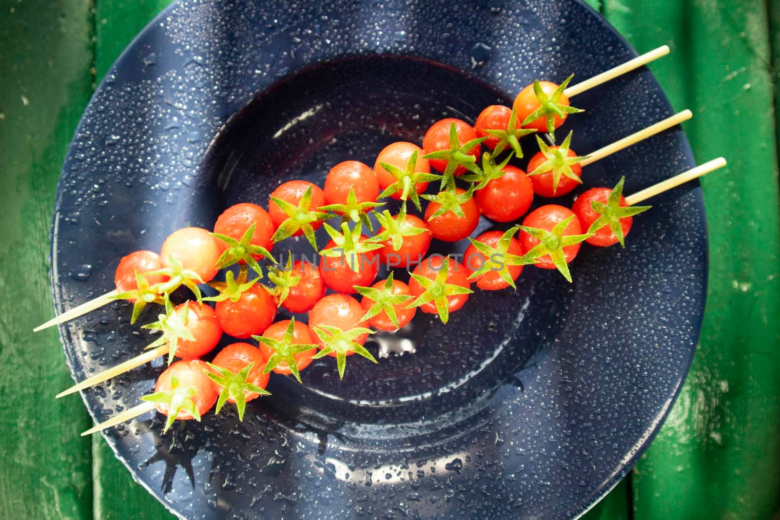 Photographic representation of three skewers made with red and tasty cherry tomatoes from Pachino, Sicily Italy placed over blue plate. 