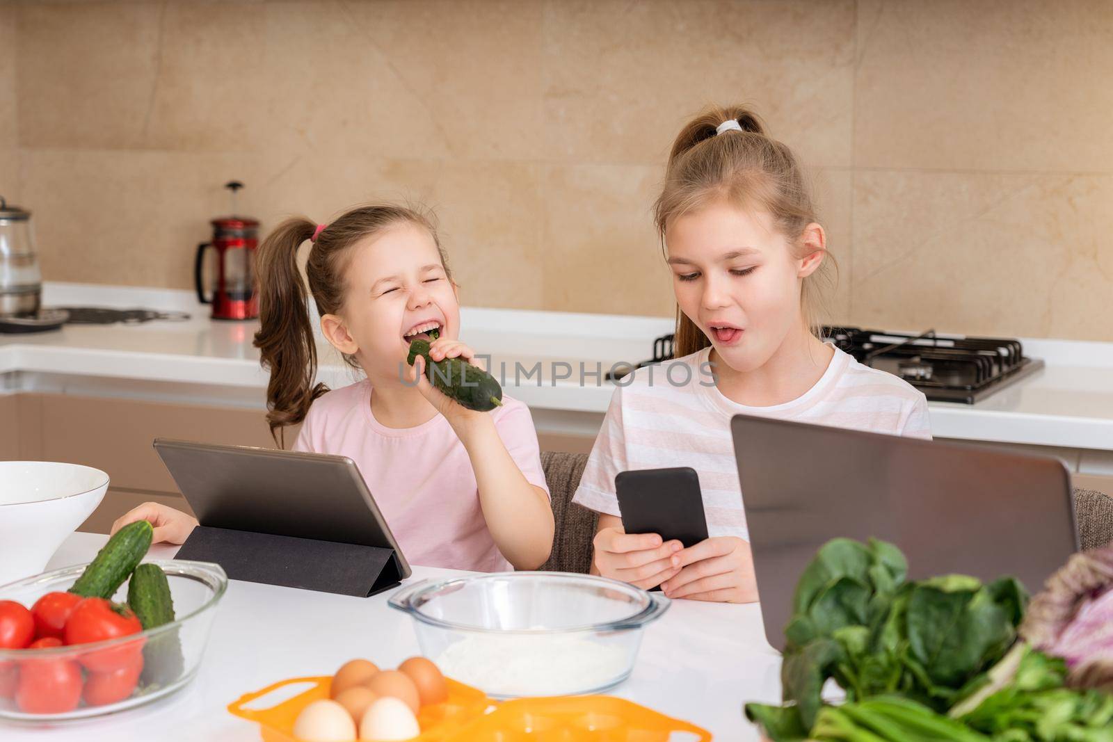Little girl eating cucumber and watching cartoons on a tablet at table in kitchen