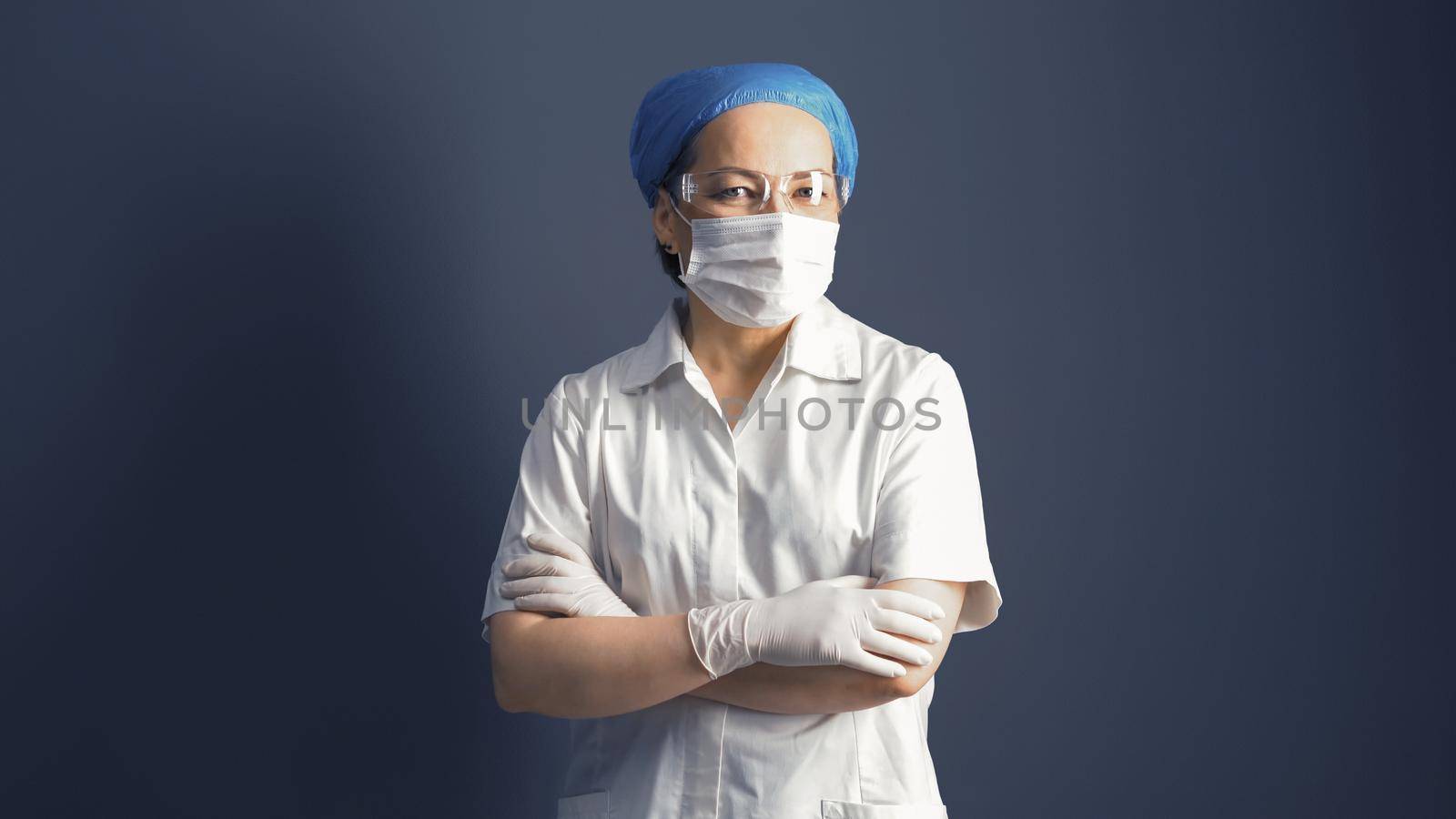 Female doctor looks at camera crossed arms. Female medic in mask and white uniform, portrait on gray background by LipikStockMedia