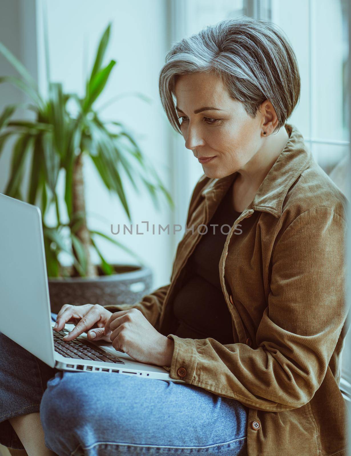 Elegant Mature woman works laptop sitting on sill of window. Side view of modern silver haired Lady in casual typing on computer keyboard. 