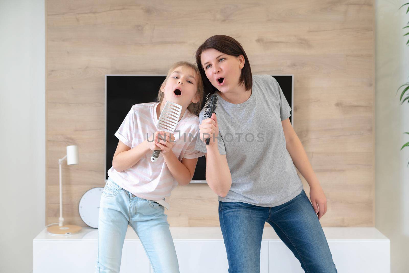 Happy family young adult mother and cute teen daughter having fun singing karaoke song in hairbrushes. mother laughing enjoying funny lifestyle activity with teenage girl at home together. by Mariakray