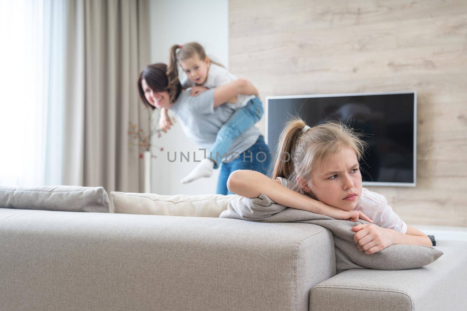 Preteenage sad girl sitting on a couch while mother having fun with sister, jealousy concept by Mariakray