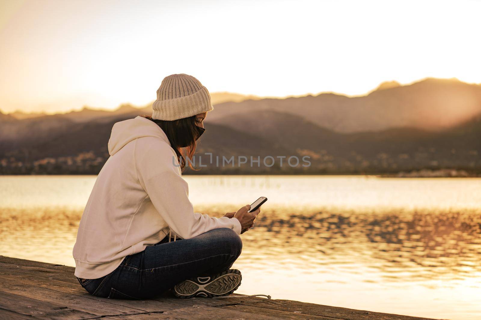 Unrecognizable young woman in white wool hat sitting on a pier alone wearing protective face mask using smartphone to keep in touch with friends and family due to Coronavirus social distancing