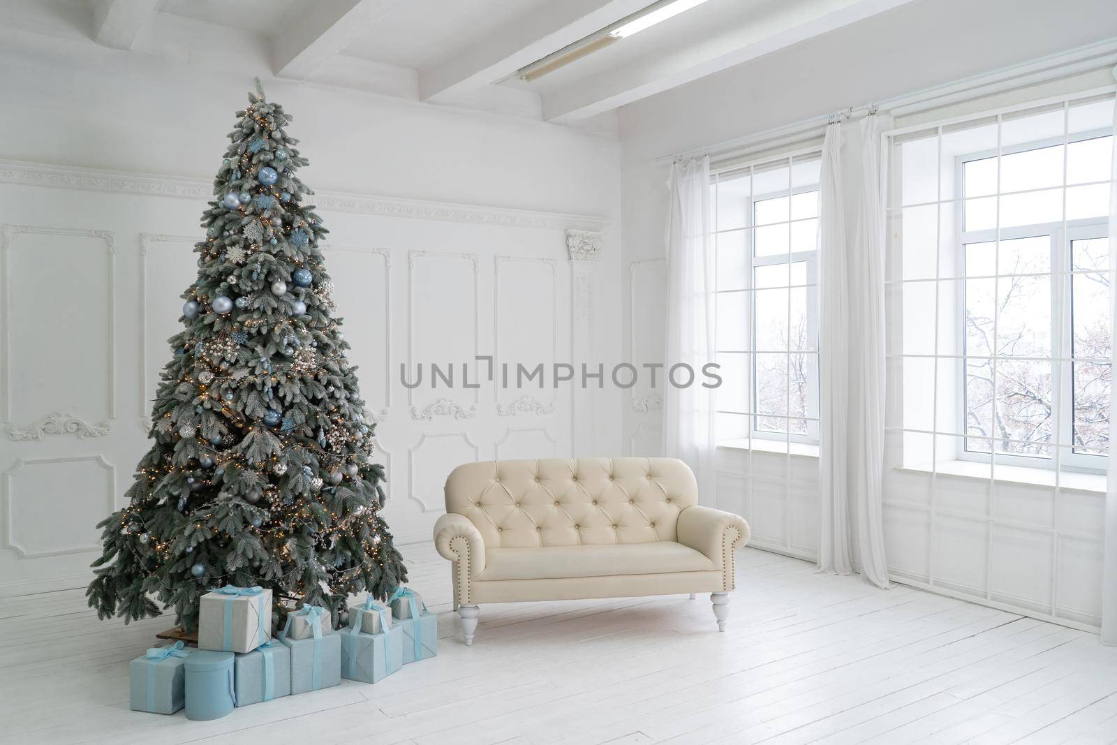 New Year studio interior. Christmas tree is decorated with balloons, with Christmas gifts under it. White sofa stands under the window and Christmas tree. High quality photo.