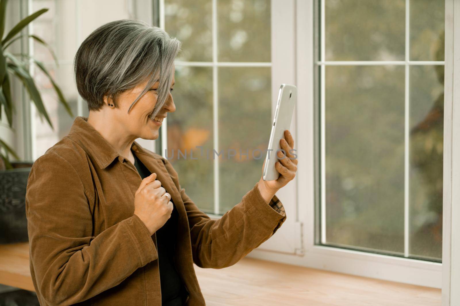 Pretty mature woman communicates online on digital tablet standing near window. Smiling woman connecting with friends by internet by LipikStockMedia