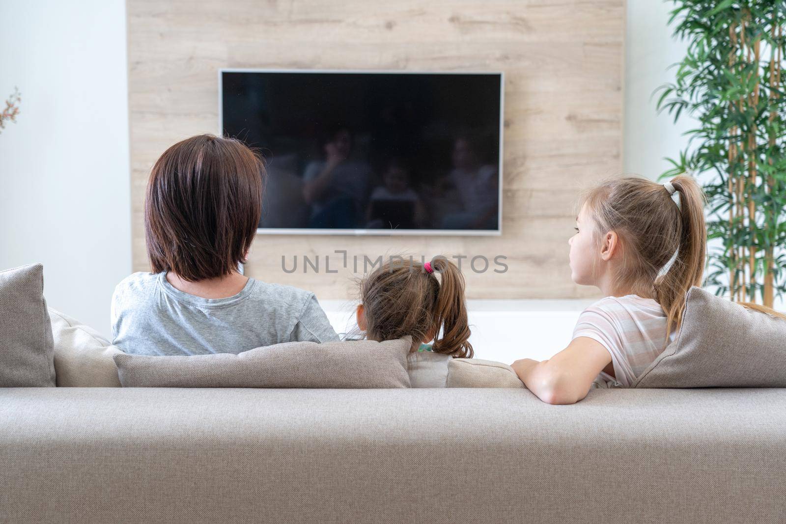 happy mother with her two cute daughters watching tv at home. Happy family relaxing on the coach. by Mariakray