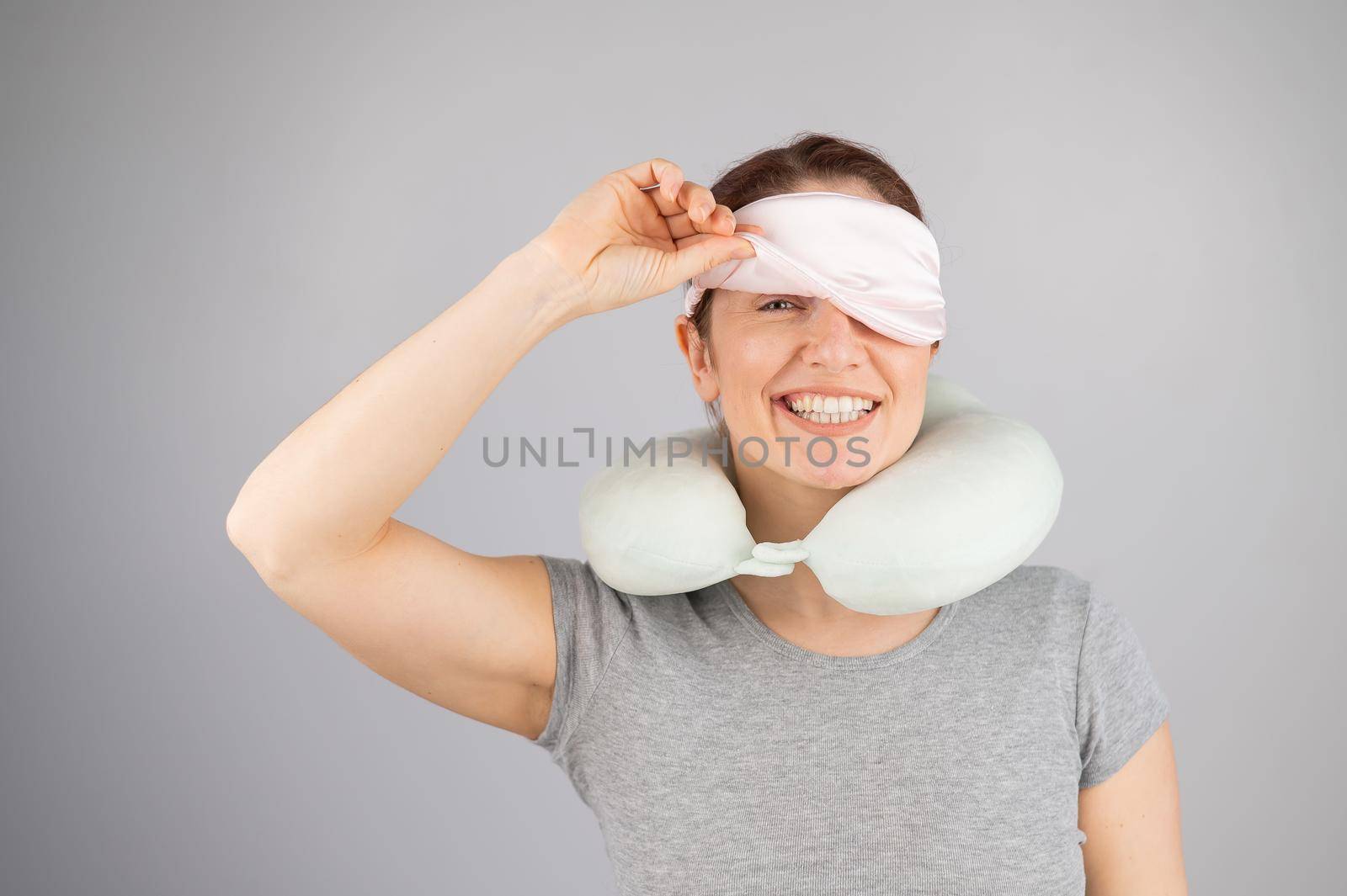 Smiling caucasian woman with a travel pillow takes off her sleep mask on a white background