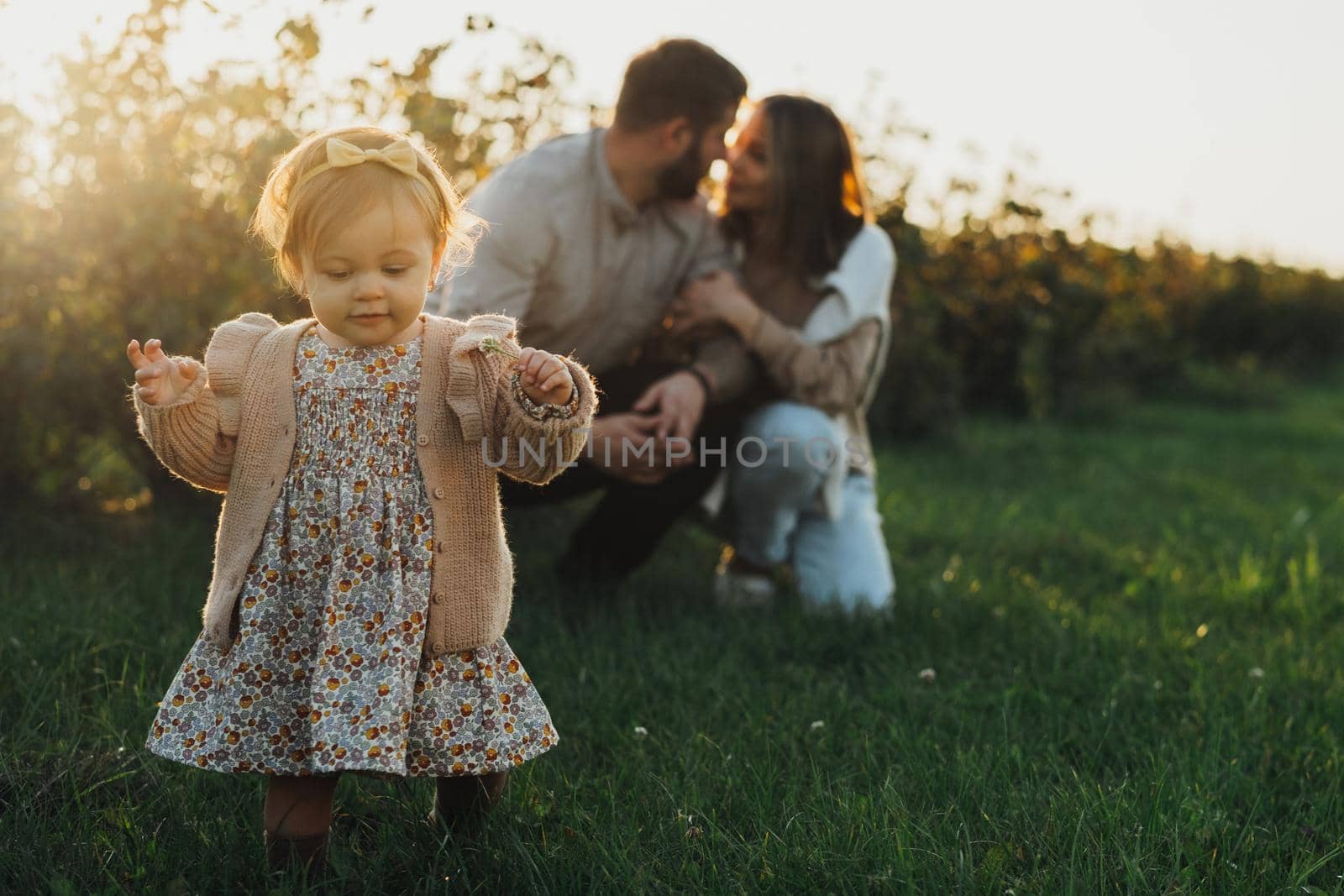 Happy Young Family with Baby Daughter Outdoors at Sunset, Little Girl Making First Steps While Her Mother and Father Hugging on Background