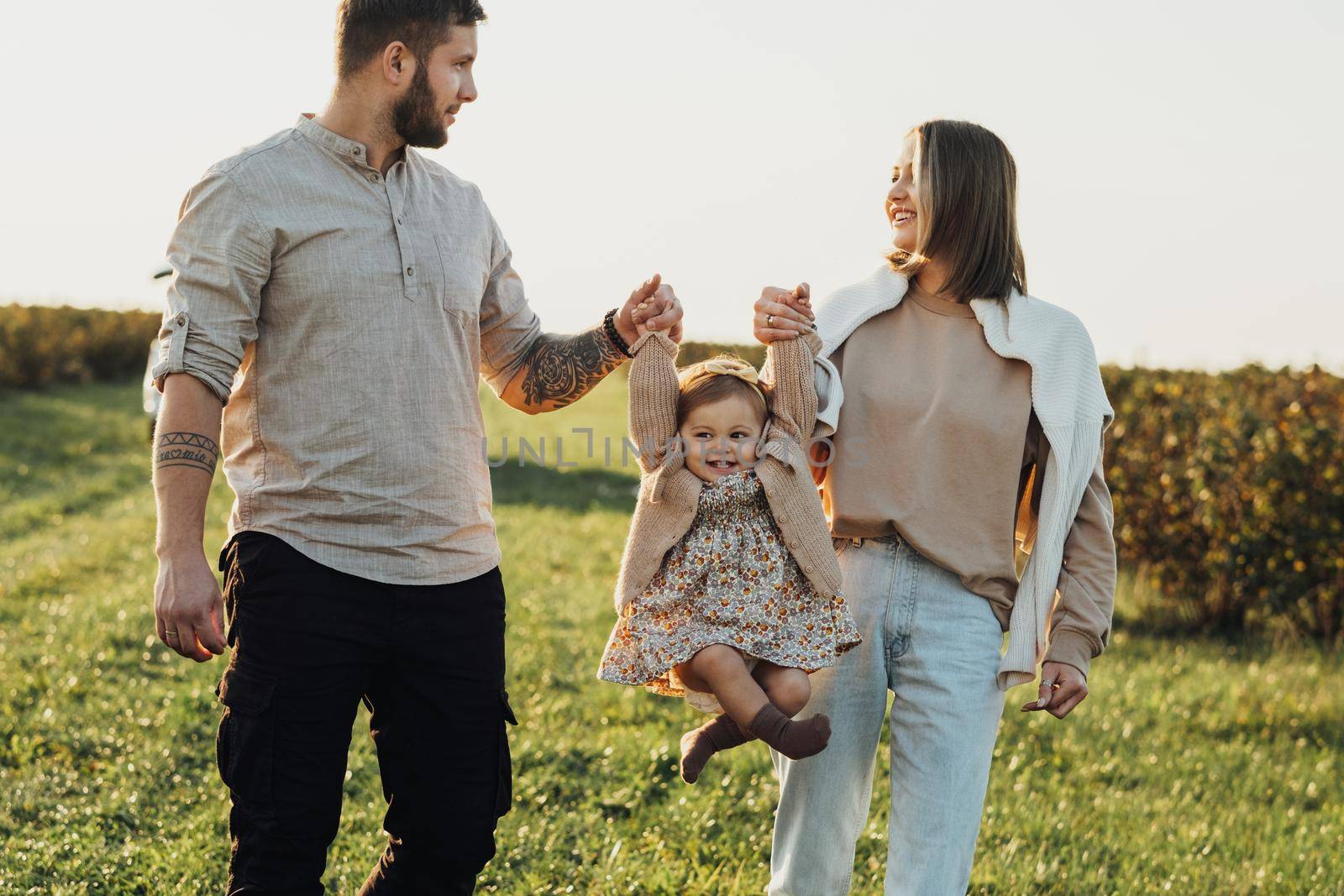 Happy Young Family Outdoors, Mother and Father Lifting Their Baby Daughter by Hands at Sunset in the Field by Romvy