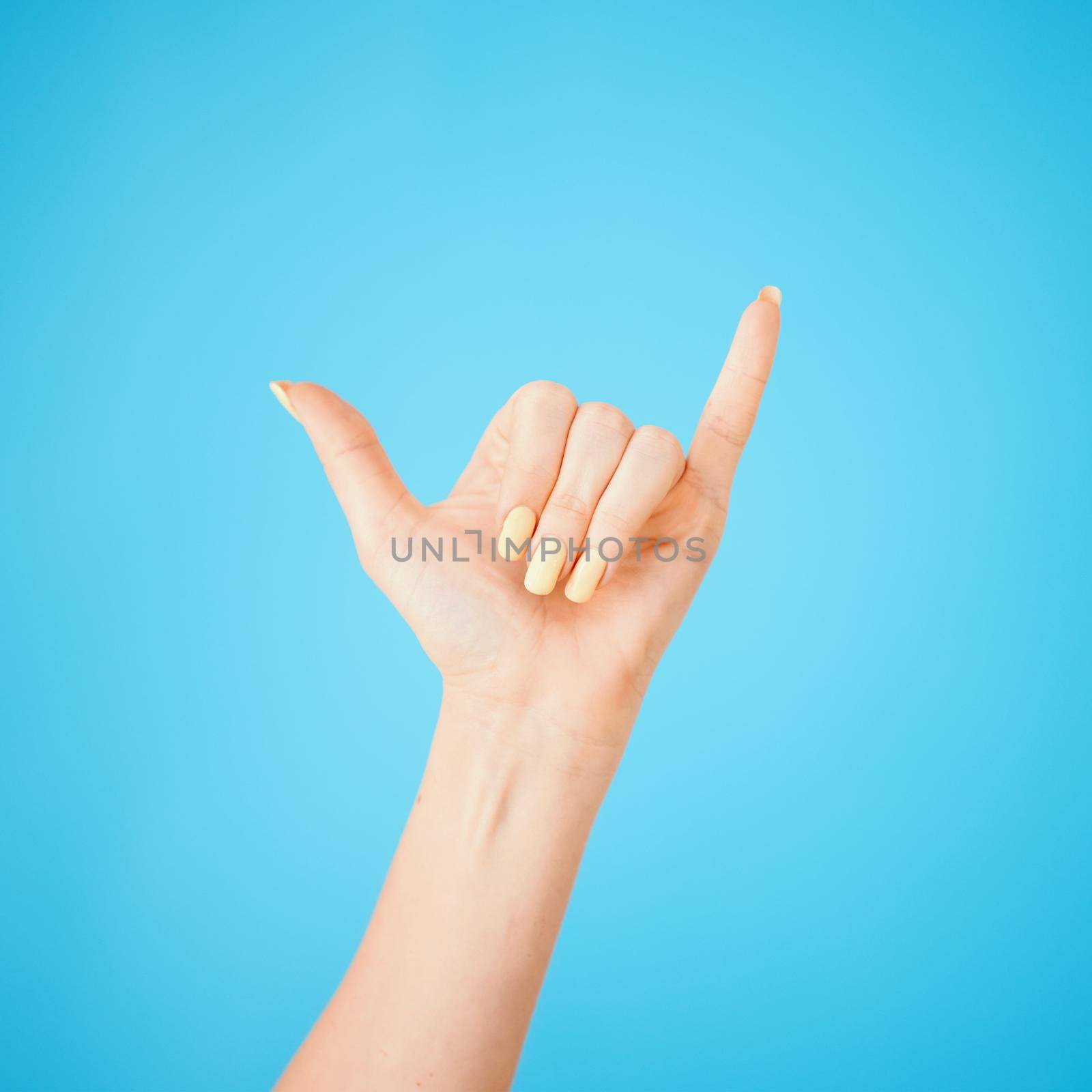 Hey, whats up. Studio shot of an unrecognisable woman showing a shaka hand sign against a blue background. by YuriArcurs