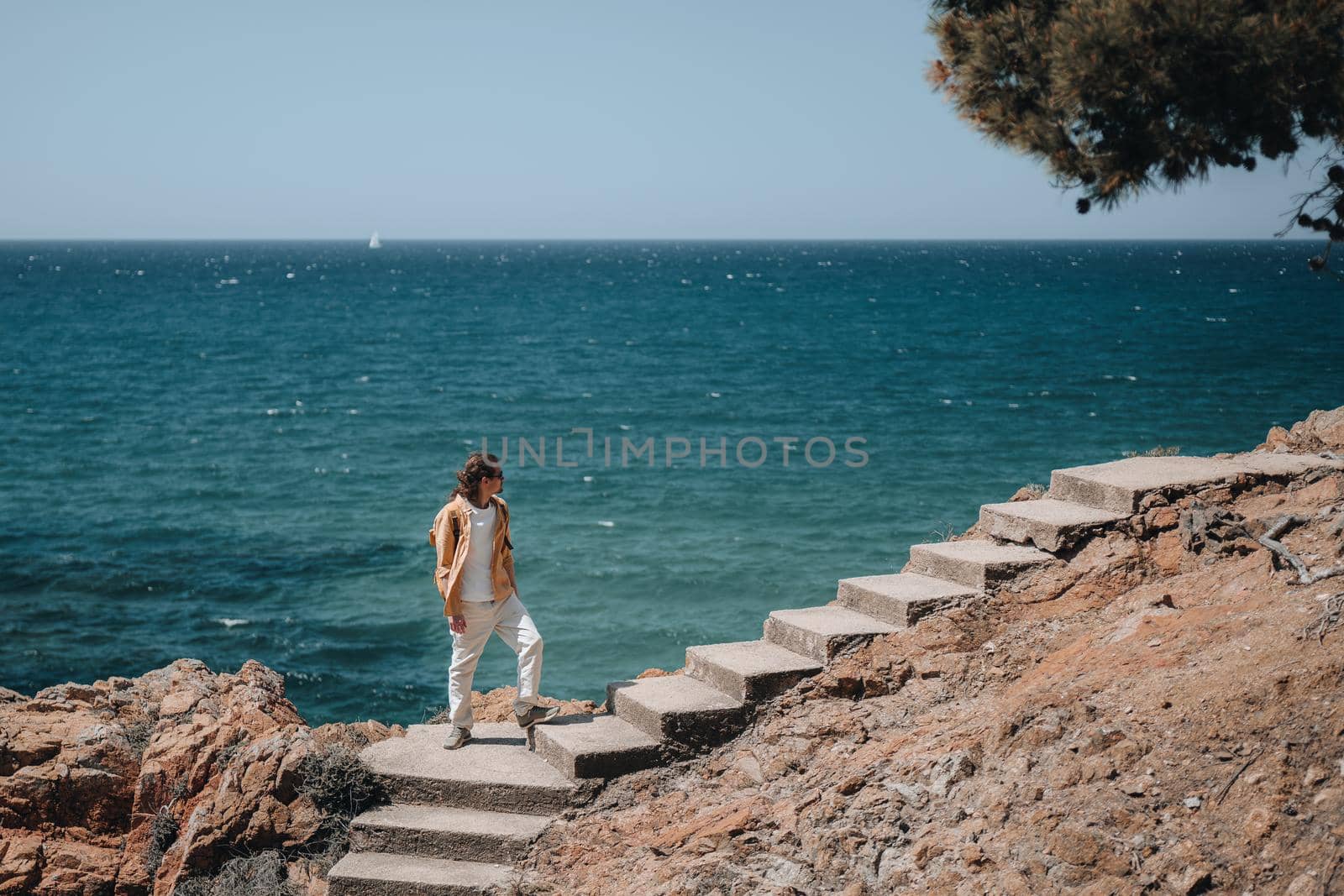 A traveler with long hair and bright closes stands on stairs and looking to the side with blue sea background. Copy space
