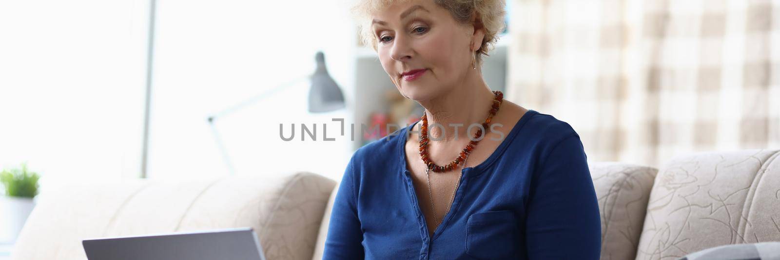 Portrait of senior woman learn how to use modern laptop device at home on sofa. Aging female explore new technology on pastime. Education, hobby concept