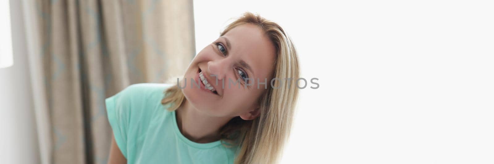 Portrait of happy smiling blonde young woman in apartment posing on curtain. Cheerful full of joy female at home. Modeling, relaxation, housewife concept