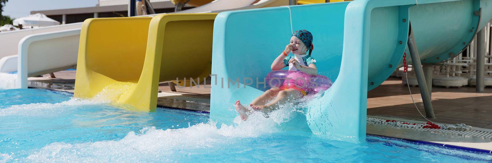 Portrait of happy little girl kid slide down hill on pink air mattress in water park. Smiling child enjoy summer vacation. Summertime, holiday, fun concept