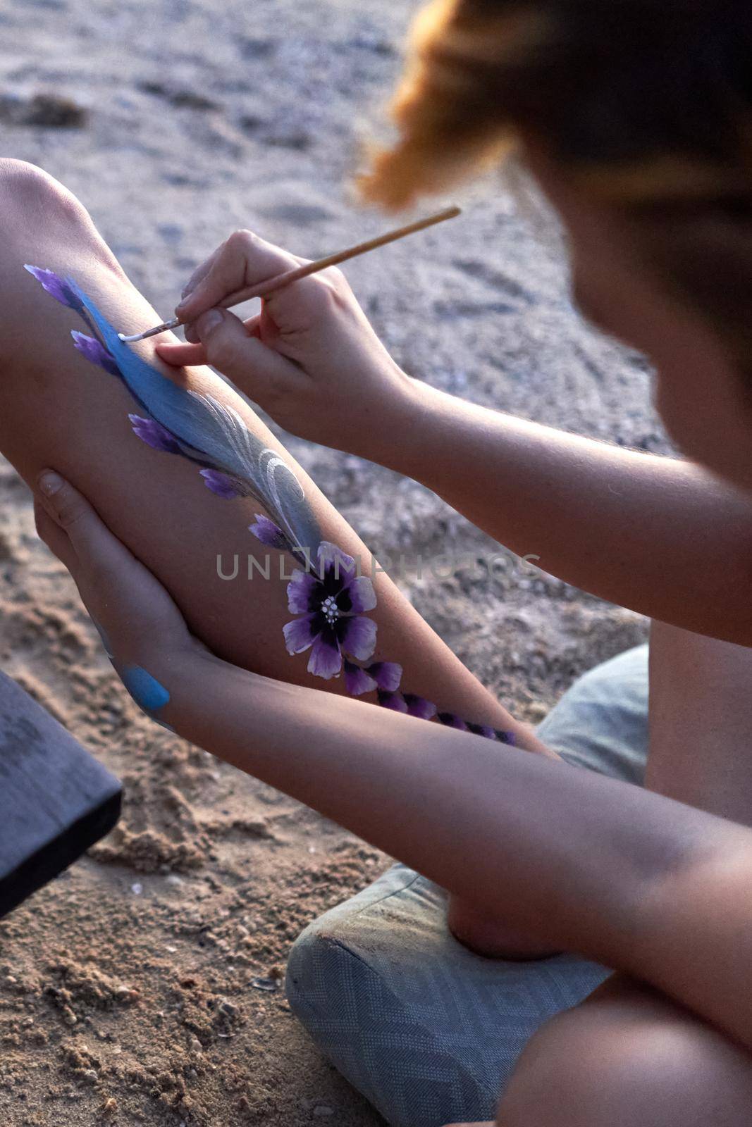 Vertical photo of a woman using a brush to paint the body of other woman on the beach