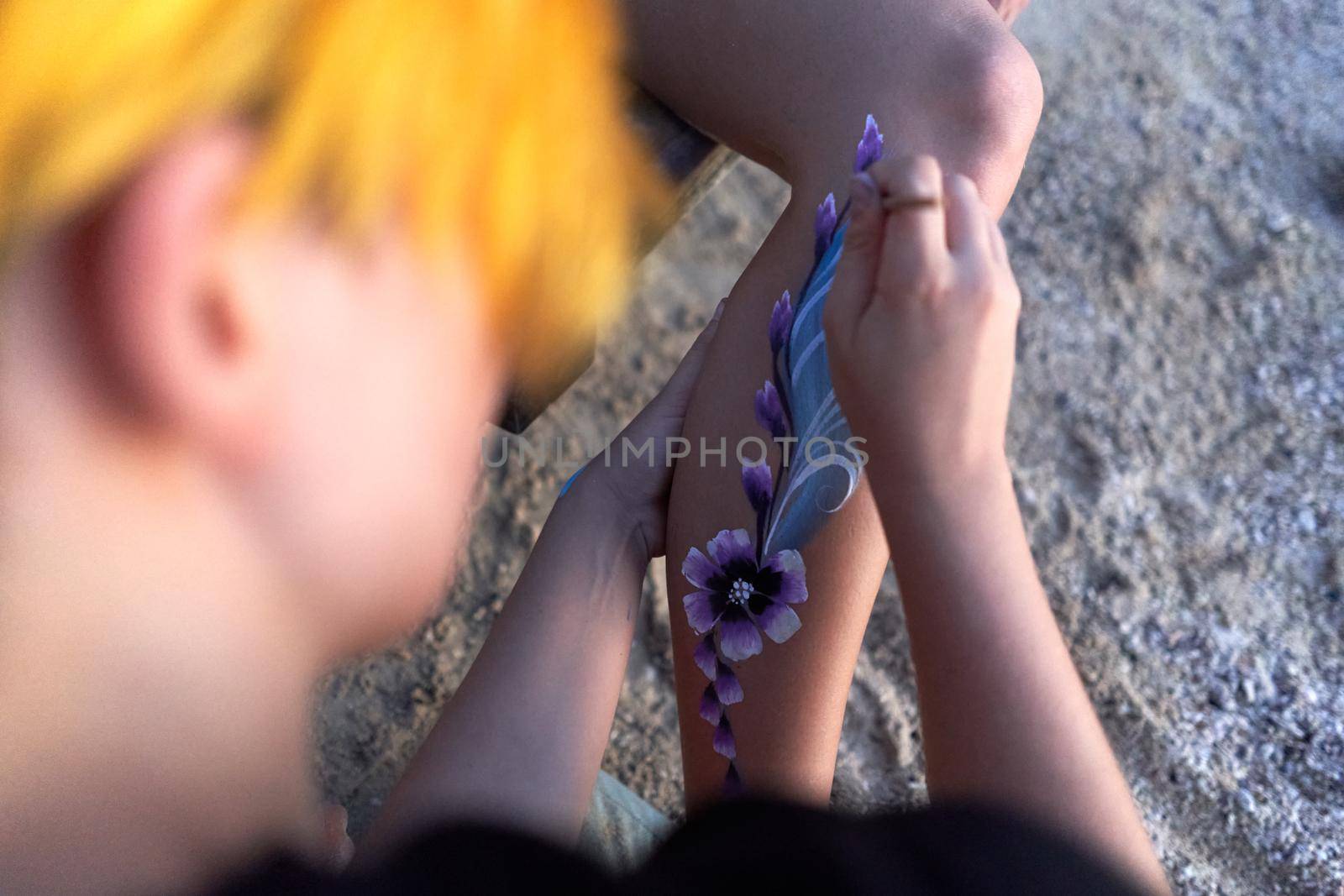 Top view and close up photo of an artist painting a flower in the leg of a woman on the beach