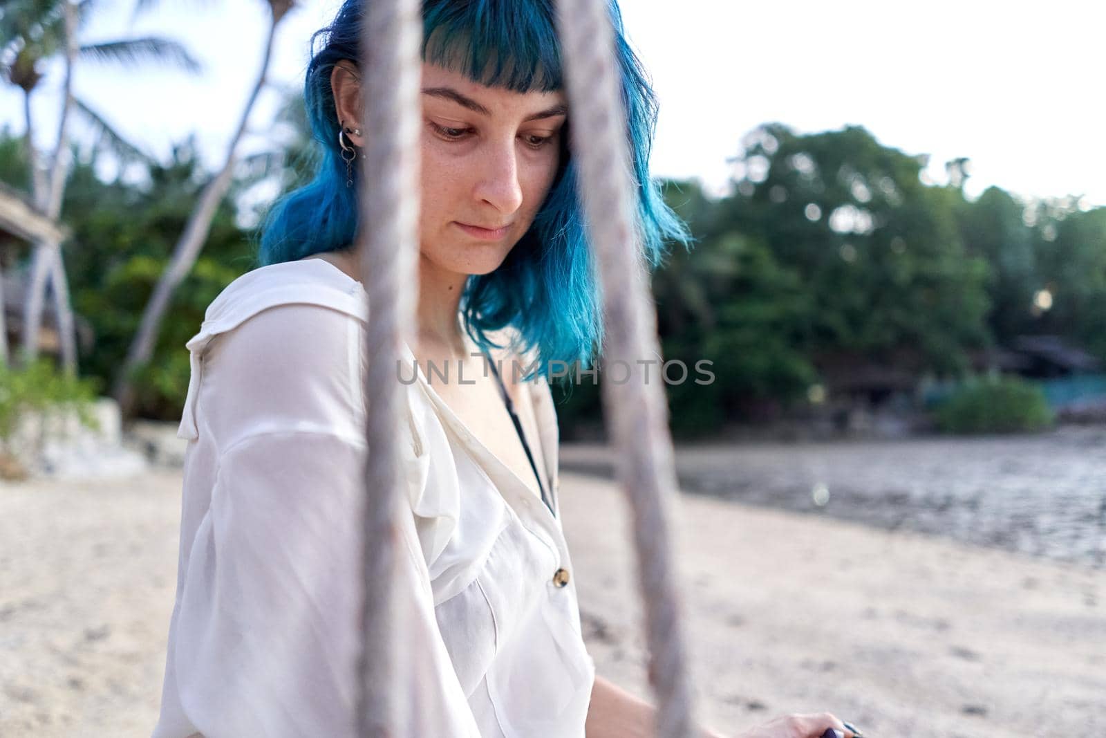 Alternative woman with blue hair sitting on a swing on a tropical beach