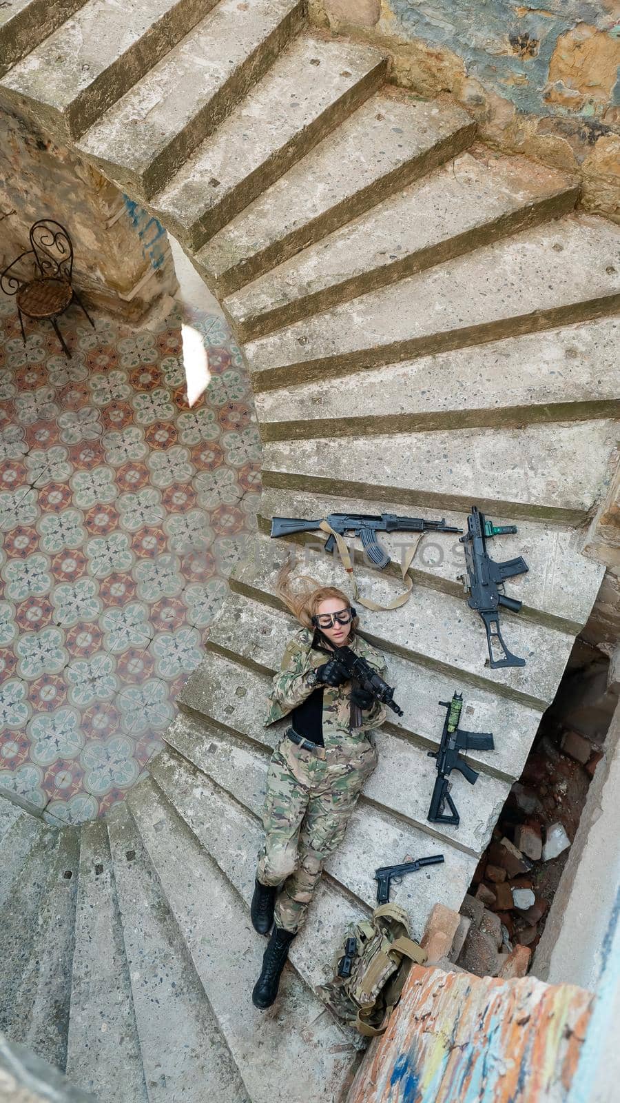 Caucasian woman in military uniform lies on the stairs of an abandoned building and holds a machine gun. View from above