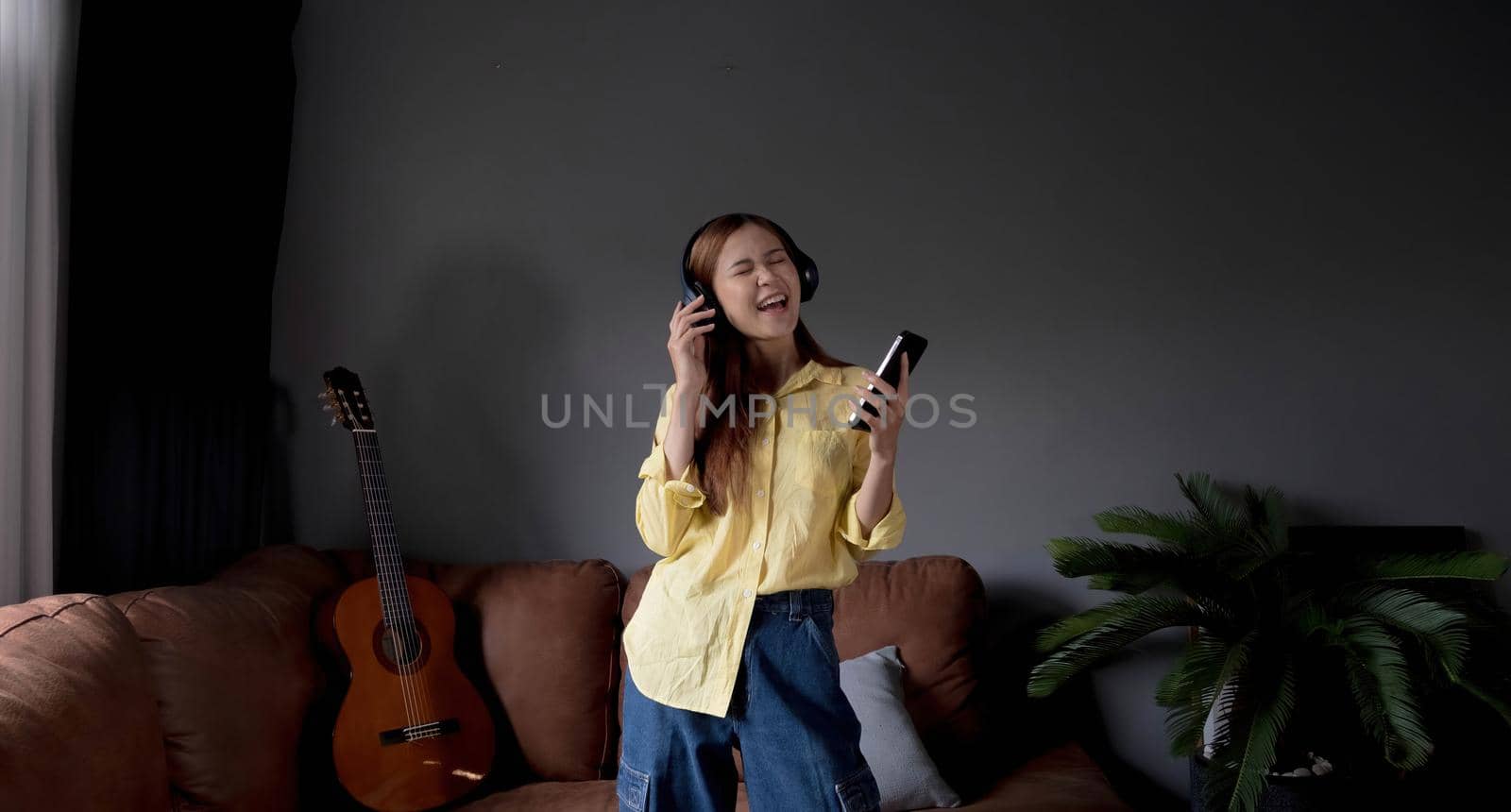 Young asian woman listening to music on couch in living room at home. Happy asia female using mobile smartphone, wearing headset by wichayada