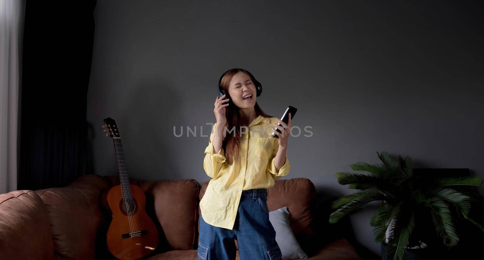 Young asian woman listening to music on couch in living room at home. Happy asia female using mobile smartphone, wearing headset