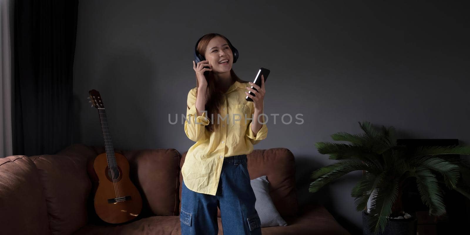 Young asian woman listening to music on couch in living room at home. Happy asia female using mobile smartphone, wearing headset and sitting on sofa.