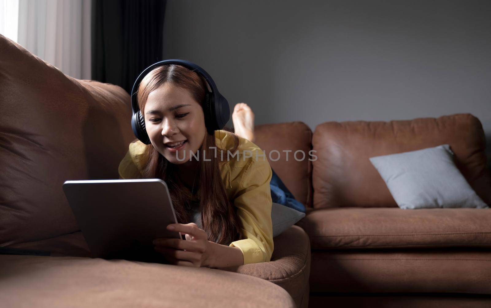 Beautiful young asian woman using laptop and wearing headphones listening to music and lying down on sofa in living room by wichayada