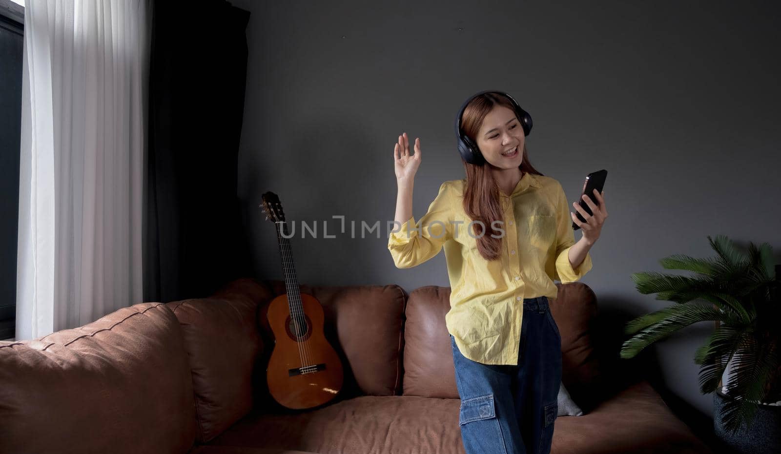 Young asian woman listening to music on couch in living room at home. Happy asia female using mobile smartphone, wearing headset