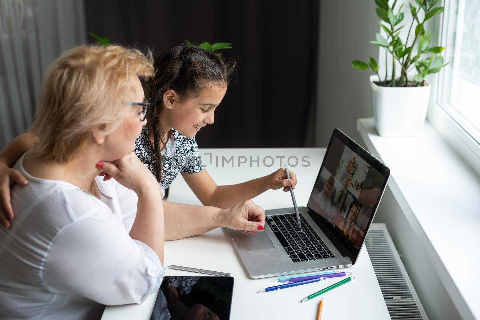 Portrait of happy grandmother and little granddaughter making video conference on pc sitting at table, waving hands at screen, greeting somebody, chatting with parents, enjoying online communication