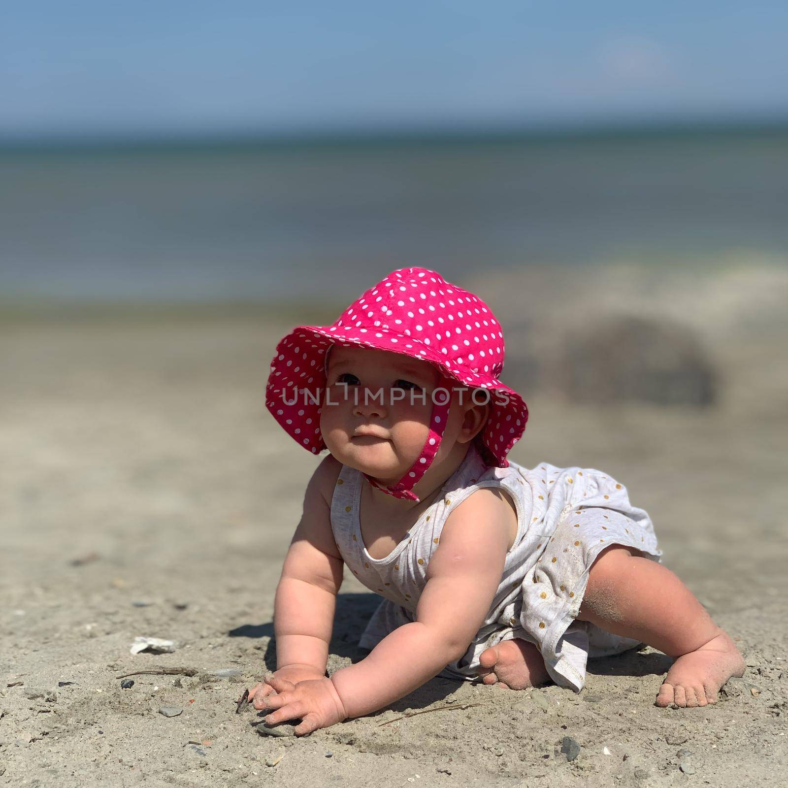 Baby girl wearing a pink summer hat crawling at the beach. Sunny day