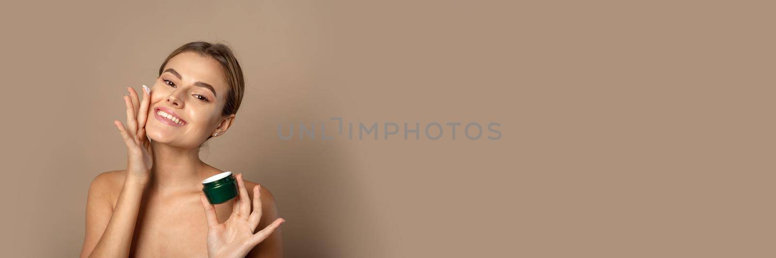 Young woman applies moisturizer to her face and smiles. Skin care concept. Web banner.