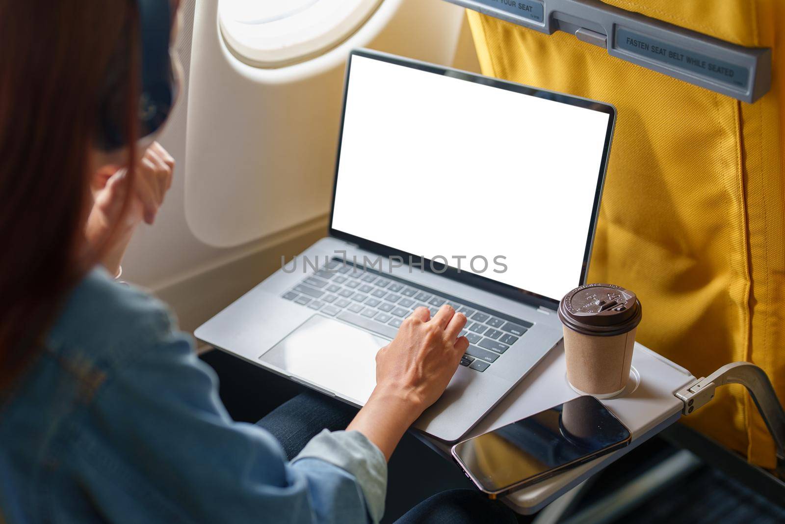 An Asian woman sits in a window seat in Economy Class using a white screen laptop computer that can use text or advertising while in flying. Travel, vacation.