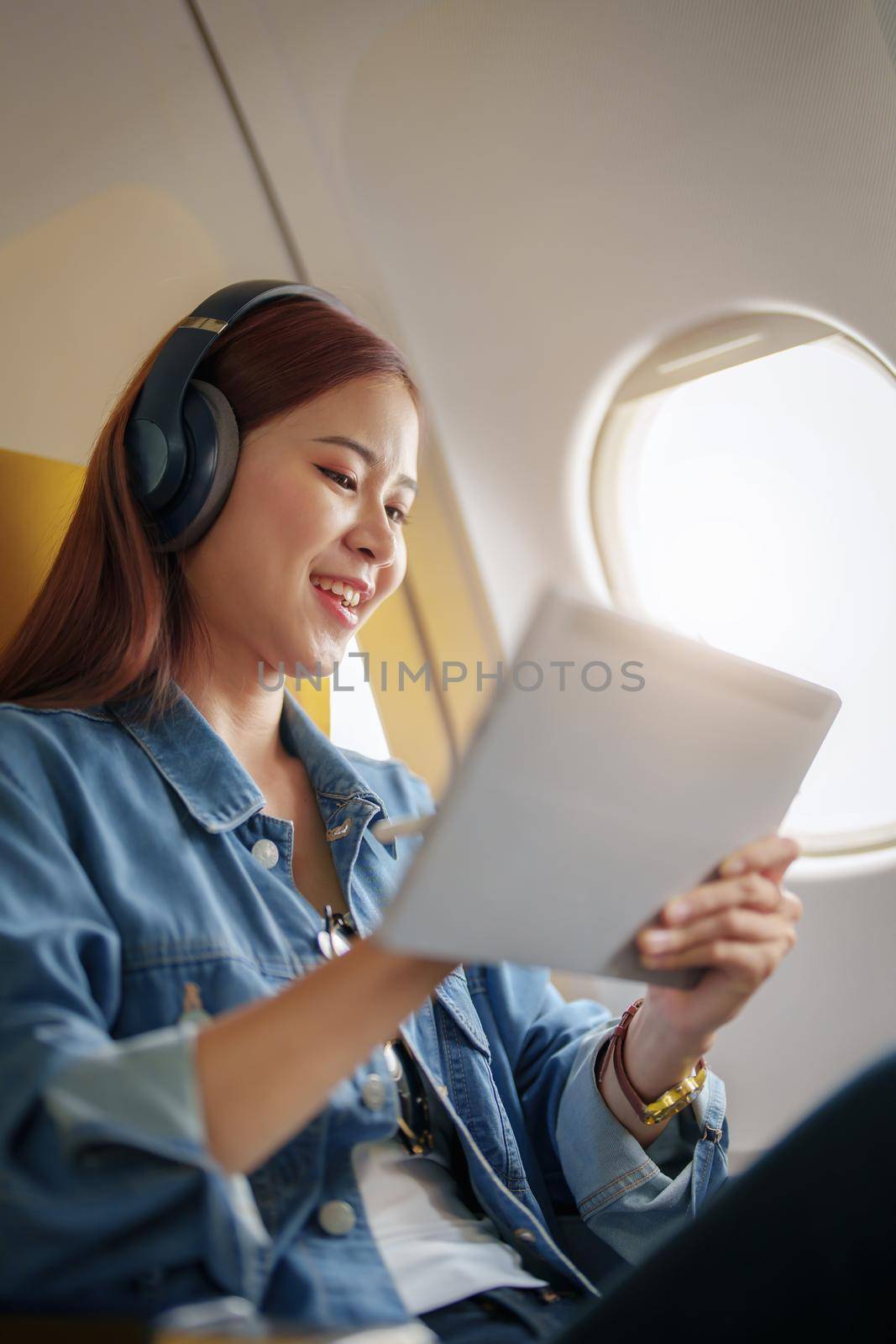 Attractive portrait of an Asian woman sitting in a window seat in economy class using tablet and listening to instrumental music during an airplane flight, travel concept, vacation, relaxation.