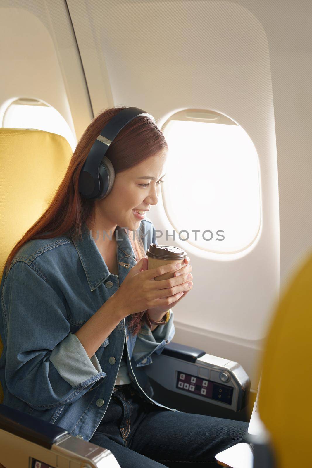 Attractive portrait of an Asian woman sitting at a window seat in economy class listening to music during a flight on a plane, travel concept, vacation, relaxation by Manastrong