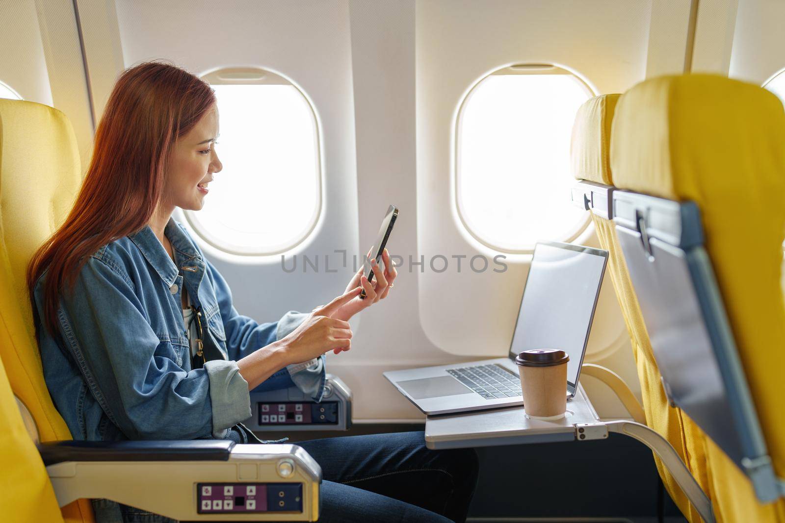 Attractive portrait of Asian woman sitting at window seat in economy class using mobile phone during inflight, travel concept, vacation, relax.