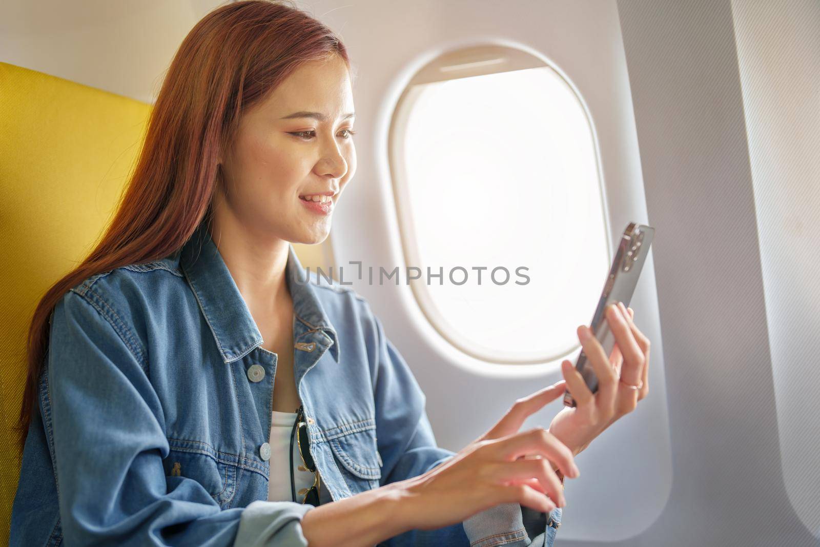 Attractive portrait of Asian woman sitting at window seat in economy class using mobile phone during inflight, travel concept, vacation, relax.