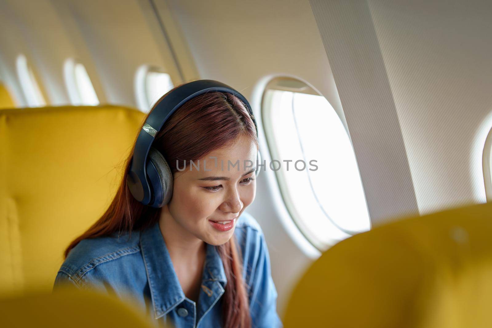 Attractive portrait of an Asian woman sitting at a window seat in economy class listening to music during a flight on a plane, travel concept, vacation, relaxation.