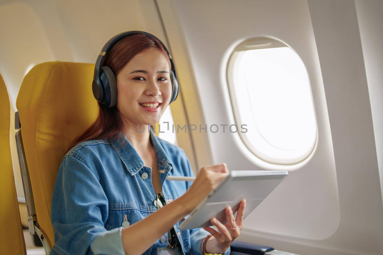 Attractive portrait of an Asian woman sitting in a window seat in economy class using tablet and listening to instrumental music during an airplane flight, travel concept, vacation, relaxation.