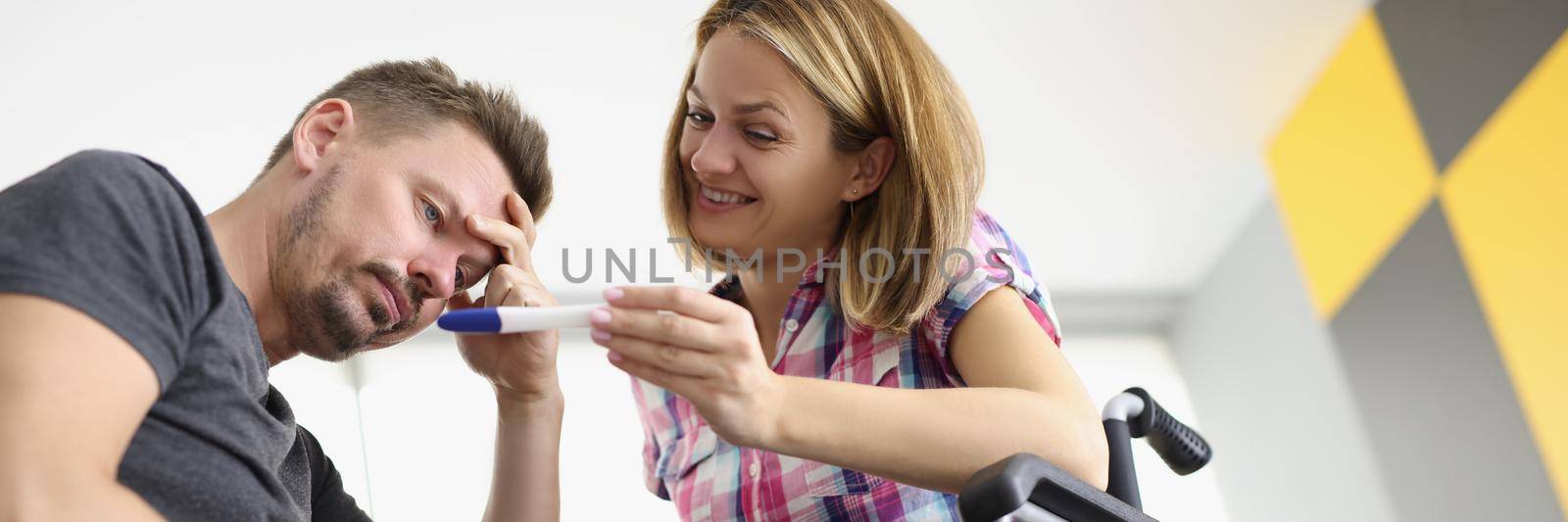 Low angle of happy female sit in wheelchair and show to husband positive pregnancy test. Upset man not happy, confused emotion. Disability, family concept