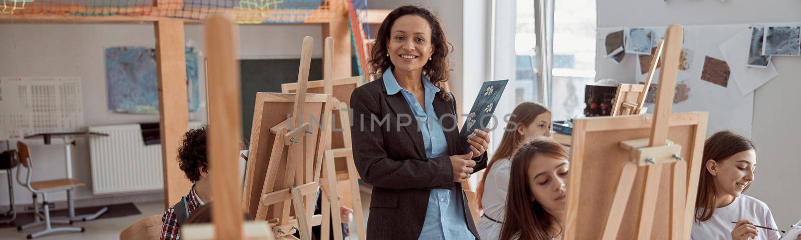 Portrait of smiling female arfican american teacher in modern classroom with kids drawing lesson in background.