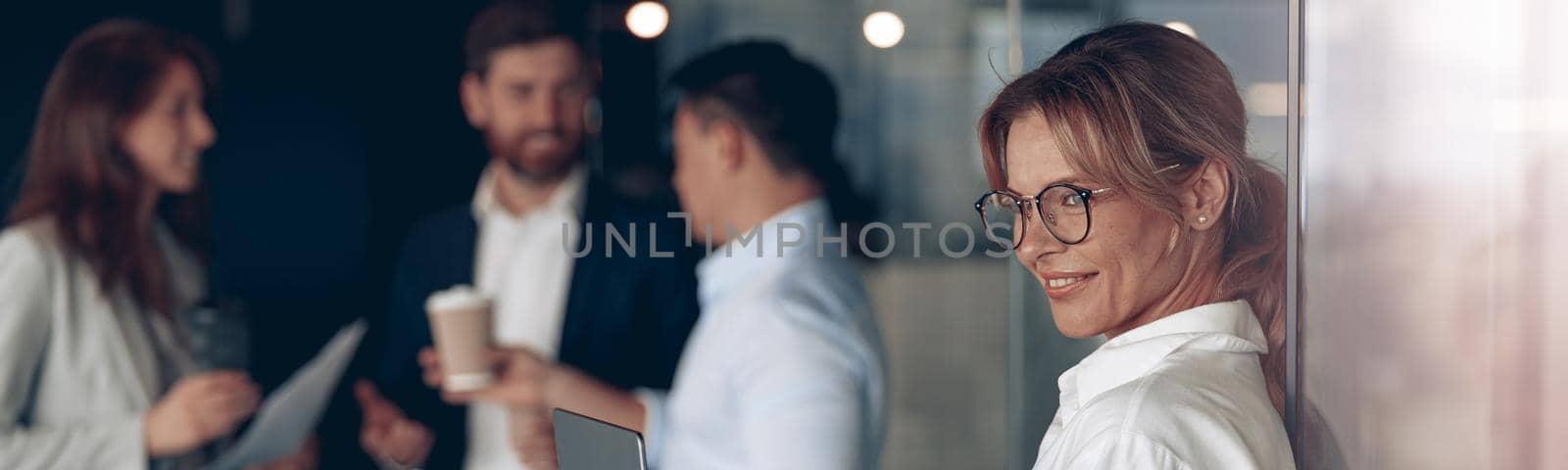 Mature smiling business lady at office with group of colleagues on background, working on laptop by Yaroslav_astakhov