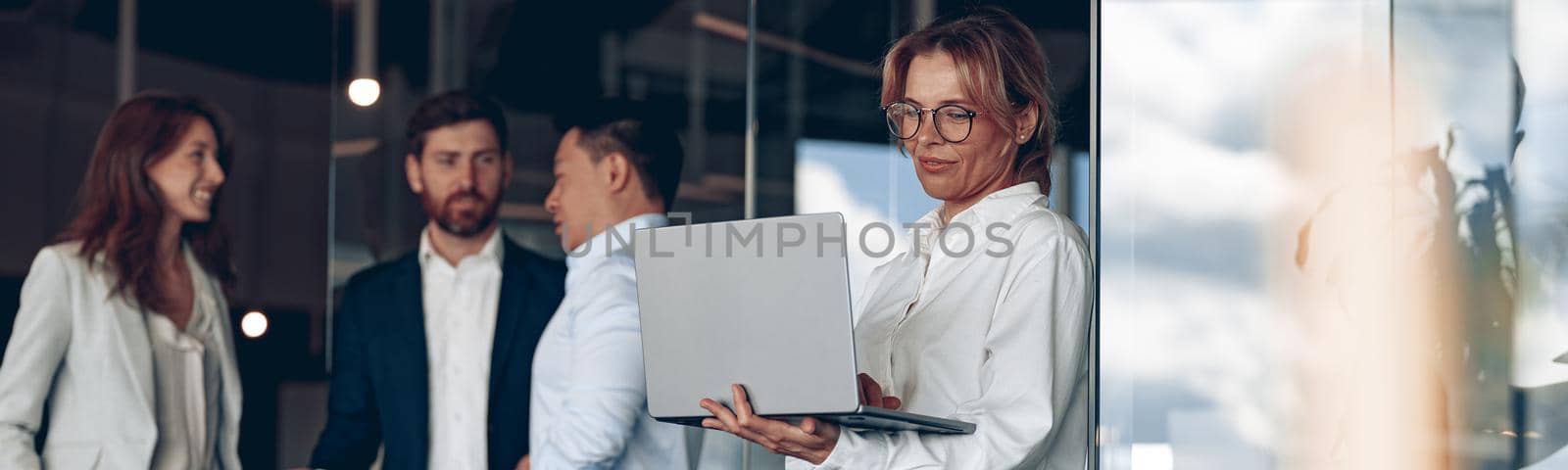 Mature confident businesswoman at office with group of colleagues on background, working on laptop.