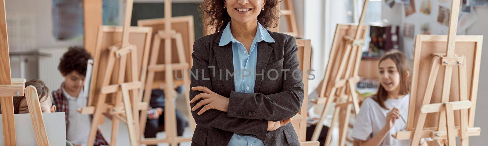 Portrait of smiling female arfican american teacher in modern classroom with kids drawing lesson in background by Yaroslav_astakhov