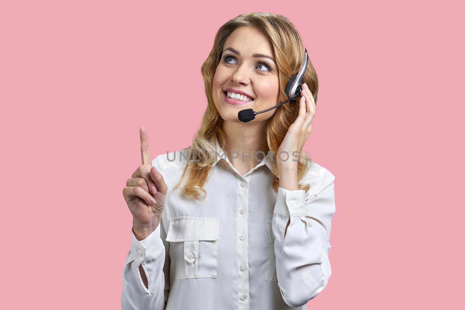 Female phone operator in headset pointing with finger upwards on pink background. Call center operator talking with a client.