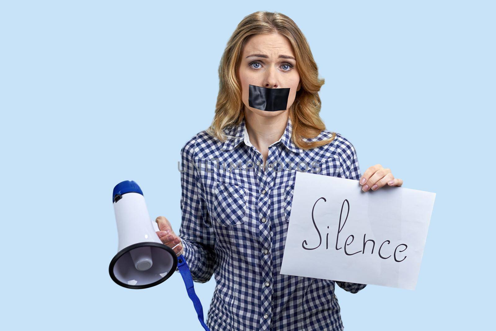 Stressed confused young woman protester with taped mouth on color background. Female activist holding megaphone and banner with inscription Silence.